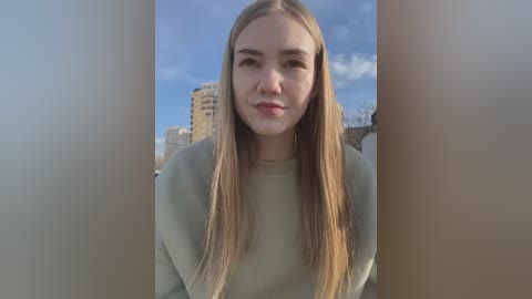 Video of a young, fair-skinned woman with long, straight blonde hair, wearing a light gray sweater, standing outdoors against a backdrop of urban buildings under a clear sky.