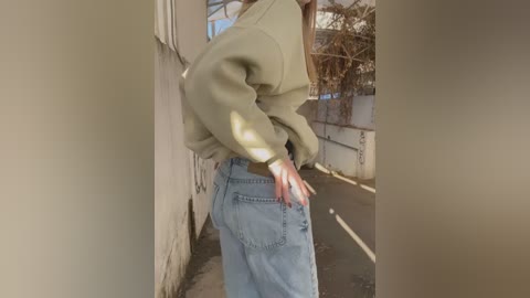 Video of a person with long hair, wearing a light green sweater and high-waisted, light blue jeans, standing in a sunlit alleyway between two beige walls.