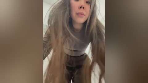 Media: Video of a woman with long, messy brown hair, wearing a grey sweater and dark pants, bending over. Background is blurry and out of focus.