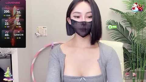 Media: Video of a young Asian woman with short black hair, wearing a gray top, black face mask, and sitting indoors with a plant in the background.