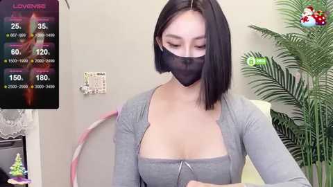 Media: Video of an Asian woman with a medium build, wearing a gray top, black face mask, and straight black hair. She stands indoors, with a green plant and a clock visible in the background.