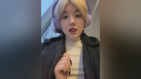 Media: Video of a young woman with blonde hair, wearing large purple headphones and a black coat over a white turtleneck, making a surprised face.