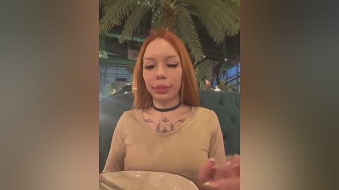 Media: A video of a young woman with long, straight red hair, wearing a beige top, black choker, and tattoos on her neck. She is sitting at a table, holding a glass, in a modern, indoor space with greenery and a palm tree.