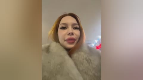 Media: Video of a young woman with fair skin, light brown hair, and a soft, pinkish lipstick, wearing a light grey fur coat, in a dimly lit room with blurred background.