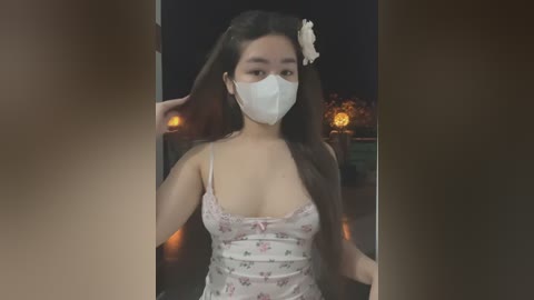 Media: Video of a young Asian woman with long dark hair, wearing a floral-patterned spaghetti-strap dress and a white face mask, standing indoors with a dimly lit background.