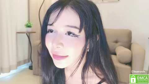 Media: Video of a young Asian woman with long, straight black hair, wearing makeup, smiling, in a beige room with a light-colored armchair.