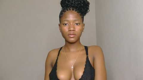 Media: Video of a young Black woman with medium brown skin and a slender physique. She has a high bun and wears a black lace bra, revealing her large breasts. She gazes directly at the camera with a neutral expression. Background is a plain, light beige wall.