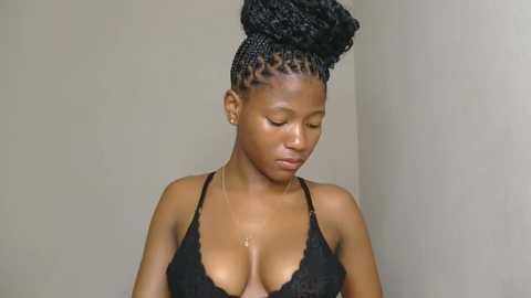 Media: Video of a dark-skinned woman with medium-sized breasts, wearing a black lace bra, her hair styled in a high, voluminous bun. She looks down thoughtfully against a plain, light-colored wall background.