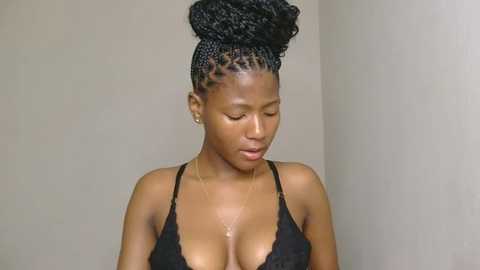 Video of a young Black woman with dark skin, wearing a black lace bralette, her hair styled in a large, intricate bun. She has a slender physique, small breasts, and is standing against a plain beige wall.