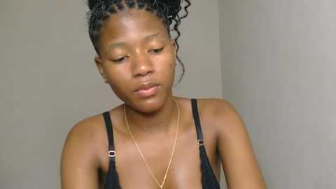 Media: Video of a young Black woman with medium-brown skin, wearing black spaghetti straps and a thin gold necklace. Her hair is styled in two tight braids. She has a neutral expression.