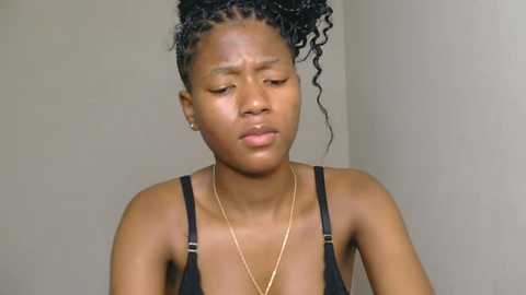 Video of a young Black woman with medium-brown skin, wearing a black tank top, braids, and a gold necklace, with a neutral expression against a plain, light-colored wall background.