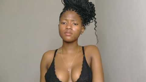 Video of a dark-skinned woman with medium-sized breasts, wearing a black lace bra and a delicate gold necklace, with curly hair styled in an updo, against a plain beige wall.