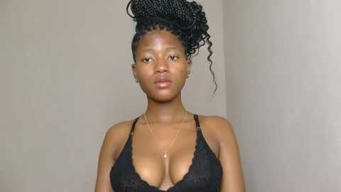 Media: Video of a dark-skinned woman with medium-sized breasts wearing a black lace bra, her hair styled in tight, curly braids. She stands against a plain beige wall, wearing a delicate gold necklace.