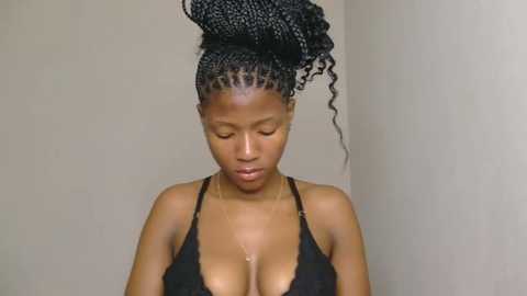 Media: Video of a dark-skinned woman with an afro hairstyle, wearing a black lace bralette, standing against a plain beige wall. Her eyes are closed, and she appears introspective.