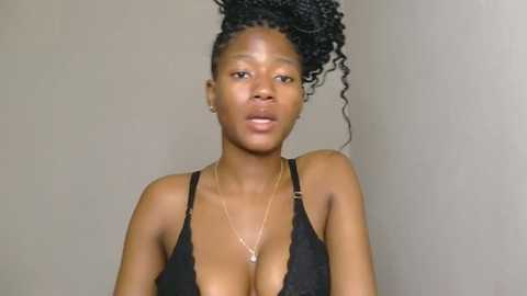 Media: Video of a Black woman with medium skin tone, wearing a black lace bralette, her hair styled in tight, black braids. She has a slender physique and medium-sized breasts. Plain background.