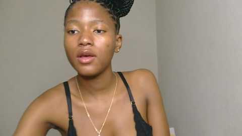 Media: Video of a young African-American woman with medium-brown skin, wearing a black bra and a thin gold necklace. Her hair is styled in braids, and she gazes thoughtfully at the camera against a plain, light-colored wall background.