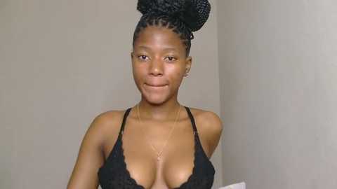 Video of a smiling Black woman with dark skin, wearing a black lace bra, her hair styled in two large braids, against a plain beige wall.