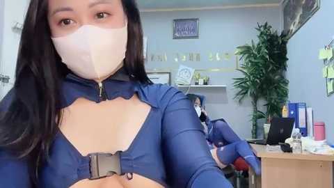 Media: Video of a woman in a blue, form-fitting superhero costume with a white mask, gloves, and belt, standing in a cluttered office with shelves, plants, and papers.