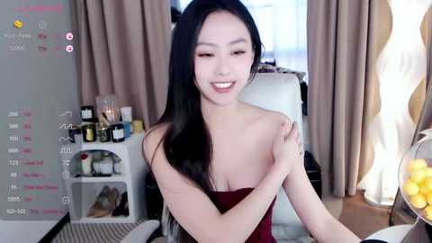 Media: Video of an East Asian woman with long black hair, fair skin, and a slender physique, smiling while seated at a table. Background includes a white chair, wine bottles, and a bowl of lemons.