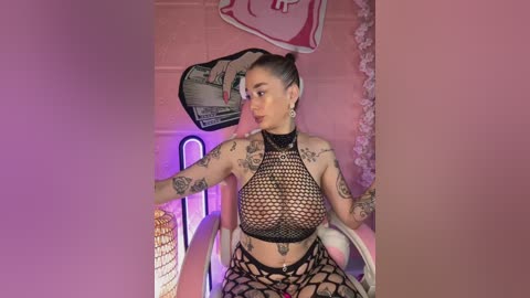 Video of a tattooed, light-skinned woman with dark hair in a bun, wearing a black fishnet crop top and matching fishnet shorts, sitting in a pink room with wall art and a neon sign.