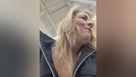 Video of a middle-aged blonde woman with wavy hair, wearing a black hoodie, tilted head, and closed eyes, standing in a brightly lit, industrial room with visible ceiling beams.