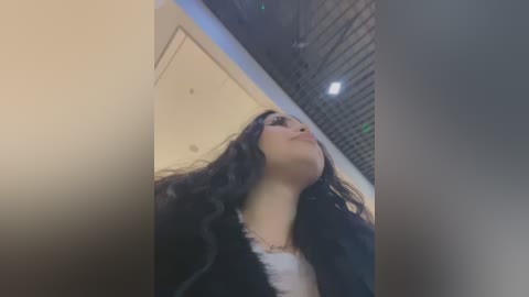 Media: Video of a young woman with long, dark hair, wearing a black jacket over a white top, looking up with a contemplative expression. Background features a modern building with metal grates and warm lighting.