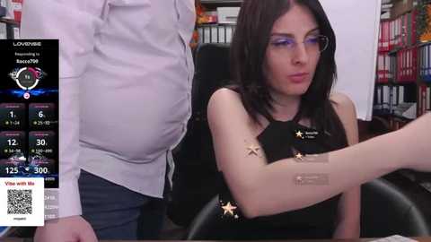 A video features a slim, pale-skinned woman with long black hair and glasses, wearing a black sleeveless top with stars, seated in an office chair. A man in a white shirt stands behind her.