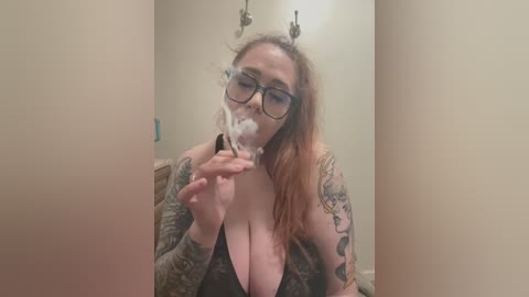 Video of a tattooed, pale-skinned woman with long, reddish-brown hair, wearing glasses and a low-cut, black top, holding a lit cigarette in a dimly lit, beige-walled room.