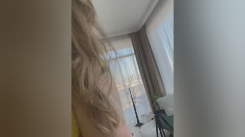 A video captures a woman with long, wavy blonde hair, wearing a yellow top, standing near a large window with sheer white curtains. A green chair and black metal stand are visible in the background.