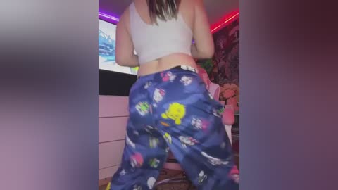 Media: A video of a woman with light skin wearing a white crop top and blue pajama pants adorned with colorful cartoon characters, standing in a cluttered room with neon lights.