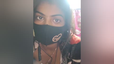 Media: Video of a woman with dark skin, wearing a black face mask and a colorful jacket, partially obscured by a blurred, bright object.