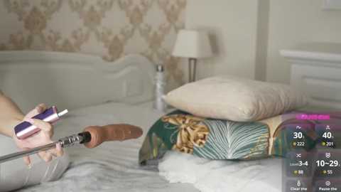 Video of a woman using a handheld vacuum cleaner on a bed with a white headboard and floral wallpaper. Bedspread has a green and beige pattern.