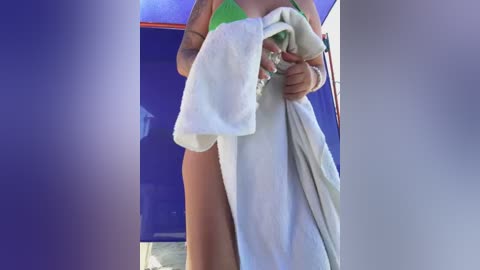 Media: Video of a woman in a green bikini, holding a white towel, partially covering her lower body, with a blue wall and blurred background.