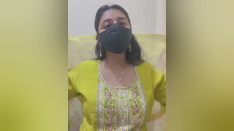 Media: Video of a South Asian woman with medium skin tone, wearing a yellow kurta with floral embroidery, black face mask, and large earrings, sitting on a beige sofa.