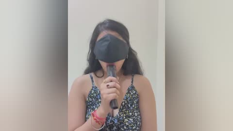 Media: Video of a woman with long black hair, wearing a black mask and a floral-patterned dress, holding a makeup brush to her face, standing in a dimly lit room with beige walls.