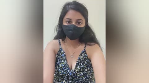 Media: Video of a young South Asian woman with long, dark hair, wearing a black face mask and floral-print sleeveless top. She has a medium complexion and a subtle smile.