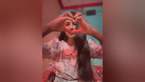 Media: Video of a young woman with long black hair, wearing a floral-patterned dress, making a heart shape with her fingers in front of her mouth. Background shows a turquoise wall and red trim.