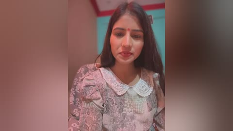 Media: Video of a young South Asian woman with long dark hair, wearing a patterned blouse with lace collar, standing in a dimly lit room with red and turquoise walls.