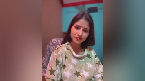 Media: Video of a young South Asian woman with medium skin tone, dark hair, wearing a star-patterned green top, seated indoors, blurred background.