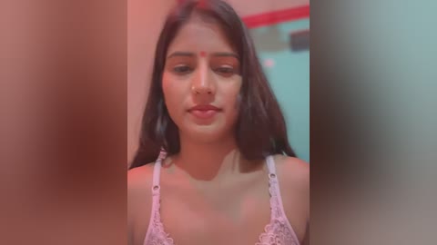 Media: Video of a young South Asian woman with medium skin tone, long dark hair, and a red bindi on her forehead, wearing a white lace bra. The background is blurred, with red and green hues.