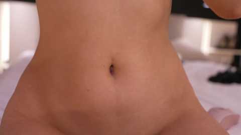 Media: Video of a nude woman's lower abdomen and navel, with a blurred bedroom background featuring white pillows and dark furniture.