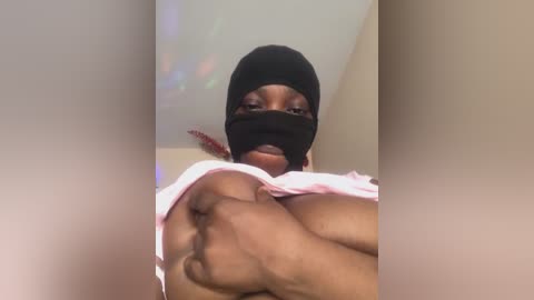 Media: A video of a young, light-skinned Black woman with a large, muscular physique, wearing a black mask and a pink top, revealing her breasts. She stands indoors against a blurred, beige wall.