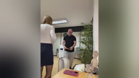 Media: Video of a woman in a white blouse and navy skirt facing a bald man in a black shirt and beige pants, standing in a modern office with a large plant and Buddha statue.