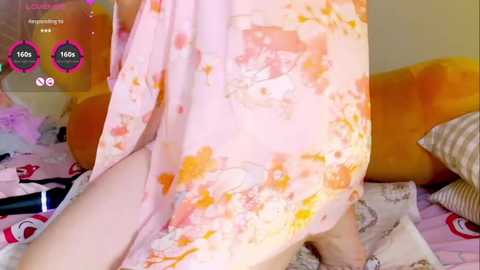 Media: A video of a person in a pink floral dress, leaning over a bed with a colorful, patterned comforter. The background features a plush, orange cushion and a pink, heart-shaped pillow.