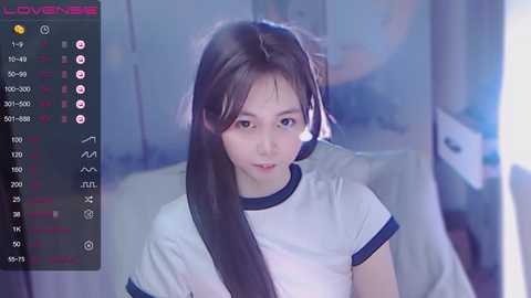 Media: A video of a young East Asian woman with long black hair, wearing a white gym t-shirt, sitting in a dimly lit room.