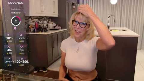 Media: Video of a blonde woman with glasses and a tight white top, adjusting her hair in a modern kitchen.