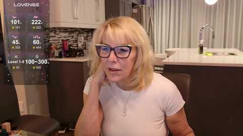 Media: Video of a middle-aged woman with short blonde hair, glasses, and a white t-shirt, seated at a modern kitchen table, with a live-stream overlay showing stats and a \"Love Is\" logo.