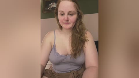 Media: Video of a fair-skinned, plus-sized woman with long, wavy brown hair, wearing a light purple spaghetti-strap tank top and brown skirt, sitting in a beige room with a framed painting on a green wall in the background.