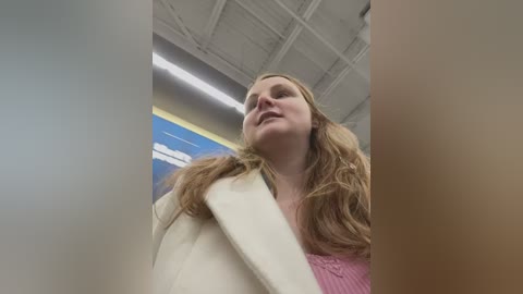 Media: Video of a blonde woman in a pink sweater, wearing a white coat, looking upward with a slight smile. Background features a blue and white ceiling and fluorescent lights.