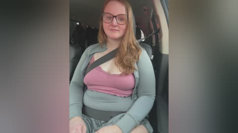 Media: Video of a plus-size white woman with long auburn hair, wearing glasses, a pink bra, and gray sweater, sitting in a car seat, with a seatbelt on.